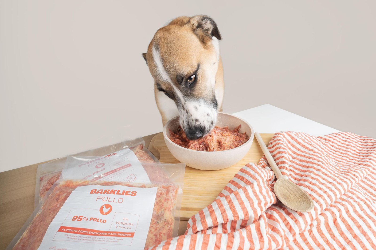 Perro disfrutando de una comida natural Barklies, hecha con 95% pollo y 5% verduras y frutas, presentada en un cuenco junto a paquetes del producto envasado y un paño a rayas naranjas.